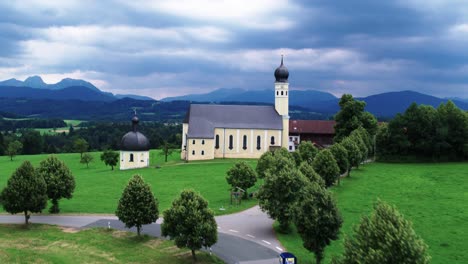 Aerial-shot-of-church-of-pilgrimage-Wilparting-with-impressive-clouds-and-flight-towards-the-Alps-with-Mount-Wendelstein-in-the-back,-Bavaria,-Germany
