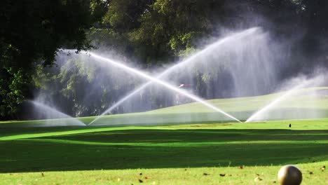Riding-lawn-mower-in-background-of-golf-course-as-sprinklers-water-the-green-grass