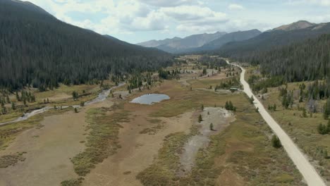 Aerial-rising-dolly-over-meadow,-road-and-river-with-pond-and-mountains-in-background-in-Colorado
