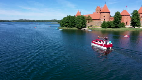 AERIAL:-Red-Boat-With-Tourists-Sailing-Past-the-Trakai-Castle-Island-and-Making-Ripples-Across-the-Surface-of-Lake