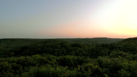 Aerial-Footage-over-forest-during-sunset,-beautiful-sky-on-the-horizon-and-green-tree-crowns-below,-natural-landscape-in-the-city,-close-to-residential-buildings