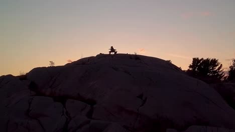 Fly-over-Inukshuk-on-Rocky-Pine-Tree-Island-at-Sunset,-Drone-Aerial-Wide-Dolly-In