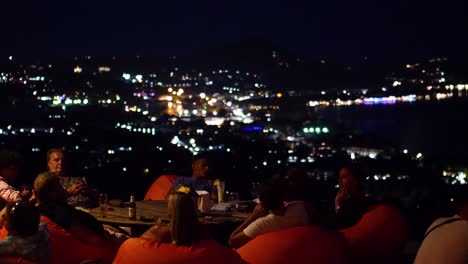 Tourists-sitting-at-the-infinity-deck-at-the-Jungle-Club-in-Samui-at-night,-with-Chaweng-town-in-the-background