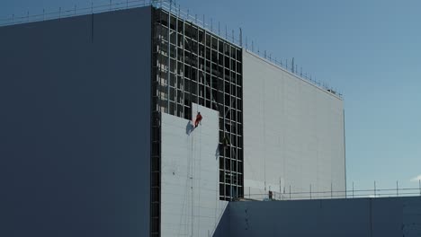 Wide-aerial-view-of-construction-workers,-abseiling-down-a-face-of-a-power-station-whilst-cladding