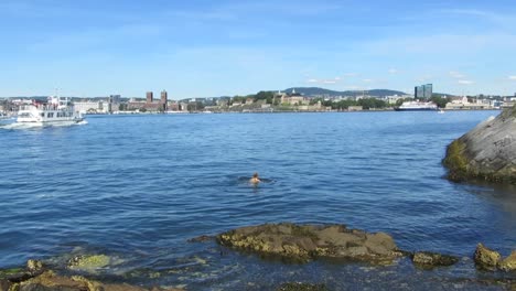 Woman-swimming-in-water-outside-Oslo,-Norway,-while-ferry-passes-by