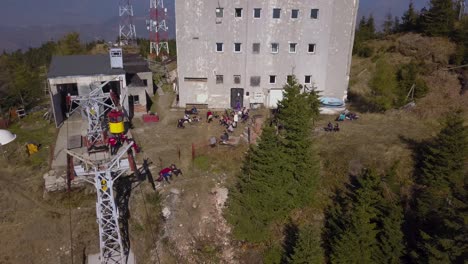 Heniu-Summit,-Romania---Hikers-Taking-A-Break-Near-A-Building-And-Towers-Surrounded-By-Green-Pine-Trees-In-The-Mountain-On-A-Sunny-Day---Aerial-Drone-Shot