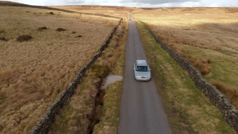 Aerial-moving-drone-shot-of-silver-car-driving-down-remote-moorland-road-in-English-countryside-on-summer-day