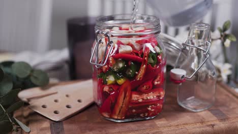 Pouring-salt-water-on-a-glass-jar-filled-with-garlic-and-mixed-chilis,-both-Habanero-and-Carolina-reaper,-standing-on-a-wooden-cutting-board-in-a-cozy-kitchen-with-flowers-and-kitchenware