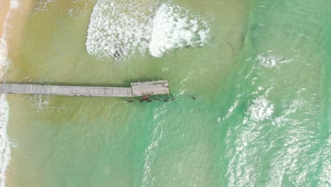 Cinematic-aerial-drone-shot-of-clear-sea-waves-crashing-onto-empty-beach-with-wooden-hand-built-pier-as-center-subject-on-Asian-island
