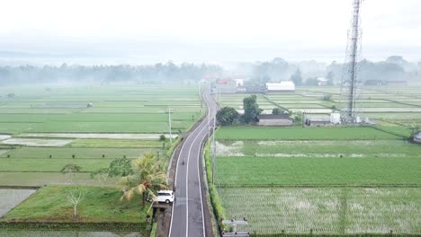 A-drone-shot-over-wet-rice-fields,-overhead-power-lines-and-a-lonely-countryside-road-in-Bali,-with-a-few-scooters-driving-in-the-early-morning-in-the-industrial-area