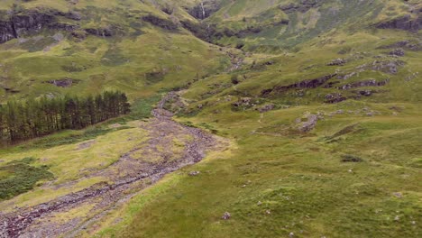 Wide-aerial-shot,-slowly-tracking-forwards-along-a-hiking-trail-and-a-rocky-mountain-stream-on-the-lush,-green-valley-hillside-of-Glen-Coe,-Scotland,-United-Kingdom,-Europe