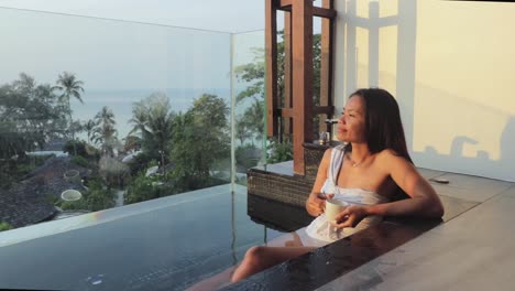 Young-girl-enjoying-a-bath-on-a-balcony-in-a-luxurious-hotel,-holding-a-cup-of-coffee-or-tea-and-admiring-a-tropical-view