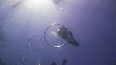 Air-bubble-ring-rising-from-scuba-diver-toward-surface-with-sun-and-scuba-divers-in-background-at-Phuket,-Thailand