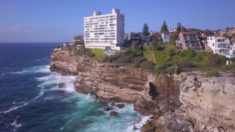 Reverse-drone-shot-of-ocean-waves-crashing-on-rocks-with-turquoise-water-and-apartment-houses-on-a-cliff-at-suburb-of-Sydney,-Diamond-bay-Australia