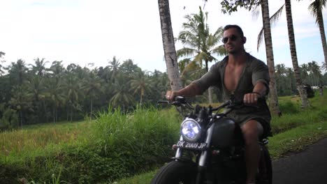 Young,-fit-and-cool-man-is-driving-on-the-countryside-roads-of-Bali-with-his-custom-motorcycle-with-beautiful-rice-fields-and-local-people-surrounding-him
