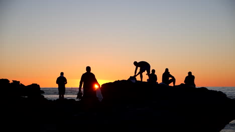Leute,-Die-Sonnenuntergang-Von-Felsen-Auf-Strandsilhouette-Beobachten