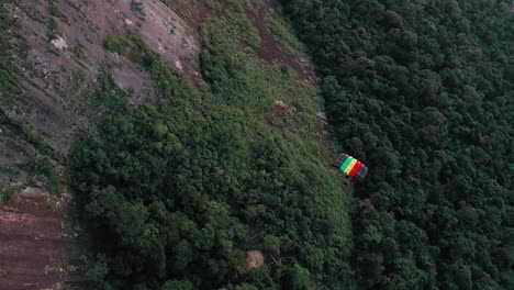 Aerial-View-Of-Parachuter-Gliding-Down-Over-Large-Rock-Face-And-Lush-Green-Forest