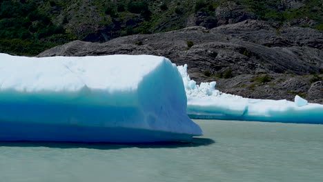 Iceberg-Azul-Iluminado-Por-El-Sol-Flotando-En-El-Lago-Gris,-Montaña-En-Segundo-Plano