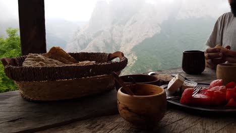 Delicious-Traditional-Breakfast-on-Handmade-Wooden-Table-in-Cottage-Terrace-with-Sliced-Fresh-Bread,-Cheese,-Tomato,-Tea,-Coffee-Hot-Drink-and-Green-Mountain-Rock-Meadow-Forest-Fog-Landscape