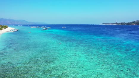 Kristallklares-Smaragdwasser-Einer-Ruhigen-Türkisfarbenen-Lagune-Mit-Booten,-Die-In-Der-Nähe-Der-Küste-Einer-Tropischen-Insel-Mit-Weißem-Strand-In-Indonesien-Schwimmen