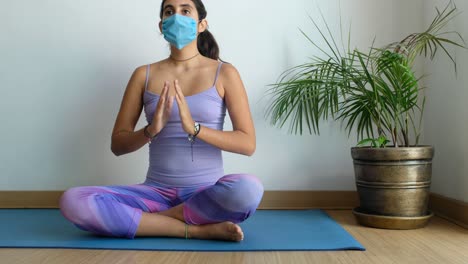 Relaxed-young-woman-in-sportswear-meditating-in-lotus-position-sitting-on-yoga-mat,-with-face-mask