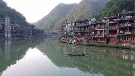 Man-sitting-and-paddling-in-an-old-wooden-boat-on-the-Tuo-river,-flowing-through-the-centre-of-Fenghuang-Old-Town