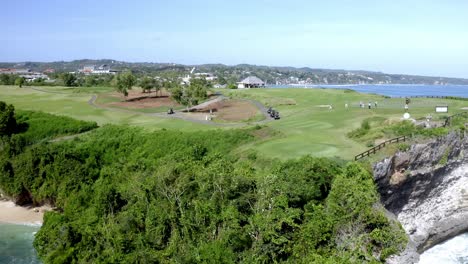 New-Kuta-Golf-Course-with-players-near-ocean-cliff-and-large-golfer-statue-at-Balangan-Beach,-Aerial-orbit-around-shot