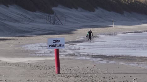 Man-In-Wetsuit-With-A-Body-Board-Running-On-The-Beach-With-Zona-Portuaria-Signage---wide-slowmo-shot