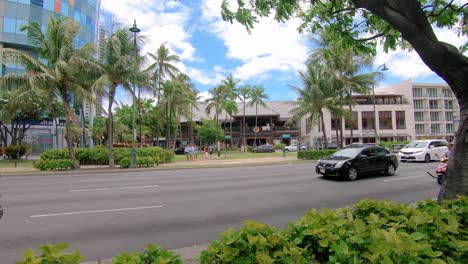 Autos,-Die-An-Einem-Sonnigen-Tag-In-Hawaii-Auf-Der-Verkehrsstraße-Mit-Luxushotels-Und-Palmen-Auf-Der-Rückseite-Vorbeifahren
