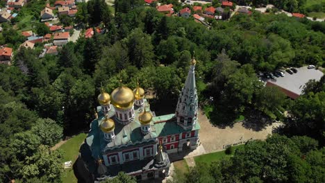 Iglesia-Conmemorativa-De-Shipka-En-Bulgaria---Hermosa-Iglesia-De-Exteriores-Coloridos-Y-Cúpulas-Doradas-Con-Exuberantes-árboles-Verdes-Circundantes---Toma-Aérea-De-Drones