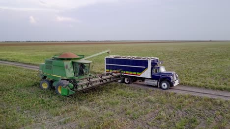 AERIAL---Combine-harvester-maneuvers-next-to-a-truck,-wide-spinning-shot