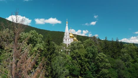 Church-Of-The-Nativity-With-Golden-Domes-In-Shipka,-Bulgaria-Surrounded-By-Dense-Green-Lush-Foliage,-Aerial-Tilt-Down-Shot