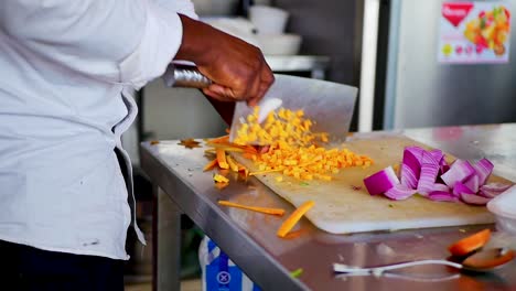 Un-Chef-Afroamericano-Cortando-Zanahorias-En-Una-Tabla-De-Cortar-Blanca-Con-Un-Cuchillo-De-Cuchilla-Grande,-Uniforme-De-Chef-De-Tiro-Estático