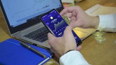 Young-Businessman-In-His-Office-Checking-Bitcoin-Chart-On-His-Phone-While-Holding-Physical-Golden-Bitcoin-In-His-Hands