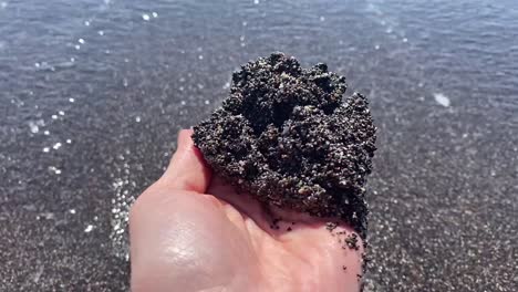 A-handful-of-black-sand-in-Vichada-Black-Sand-Beach-in-Santorini,-Greece
