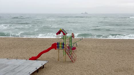 Lonely-Abandoned-Colorful-Children-Playground-on-Empty-Sea-Beach,-Ship-Moving-on-Background,-Establishing-Shot