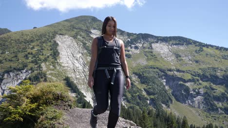 Female-hiker-with-backpack-and-hiking-boots-walks-on-a-stony-path-in-the-blazing-sun-with-mountain-range-in-the-background