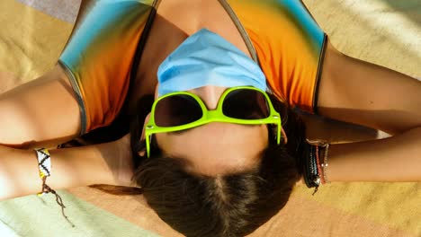 Sommer-Covid,-Mädchen-Mit-Gesichtsmaske-Setzt-Sonnenbrille-Auf-Und-Legt-Sich-Auf-Buntes-Handtuch