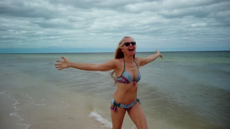 Front-shot-of-playful-blonde-mature-woman-in-sunglasses-and-bikini-running-along-the-shore-laughing-and-putting-her-arms-up-in-the-air-showing-joy