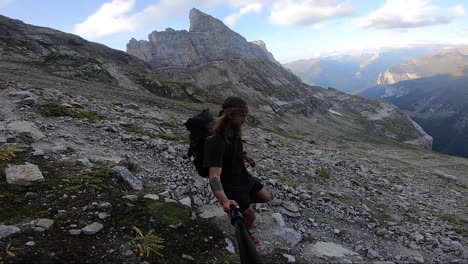 A-young,-fit-man-with-long-hair-and-a-large-backpack-is-jogging-and-running-carefully-down-a-path-in-themountains-full-of-rocks-and-stones