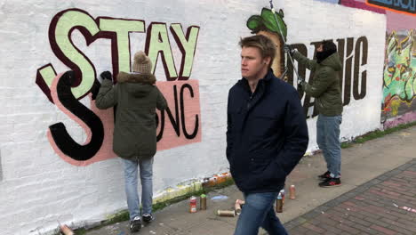 Graffiti-artists-spray-paint-a-character-wearing-a-surgical-face-mask-and-the-anti-Coronavirus-message-“stay-strong-and-don’t-panic”-on-a-wall,-as-people-pass-by