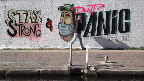 People-walk,-cycle-and-run-past-anti-Coronavirus-graffiti-art-of-a-character-wearing-a-surgical-face-mask-and-the-message-“stay-strong-and-don’t-panic”-on-a-wall-in-East-London