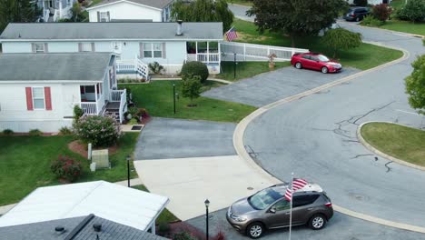 American-mobile-home-park,-USA-flags-flying-by-trailer-park-with-cars-parked-in-driveway,-summer-daytime-aerial-establishing-shot