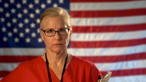 Retrato-Medio-Apretado-De-Una-Enfermera-De-Atención-Médica-Con-Portapapeles-Que-Parece-Preocupada-Y-Triste-Con-Una-Bandera-Estadounidense-Desenfocada