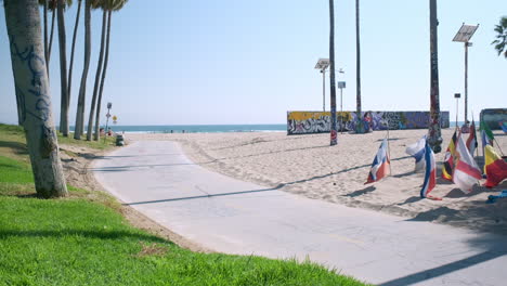 Casual-Sunday-on-Venice-Beach,-Pedestrians-and-Bikers-Leisurely-Passing-By,-Flags-and-Palm-Trees