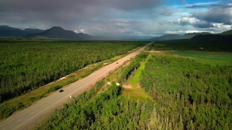 Scenic-summertime-flight-above-straight-narrow-countryside-Alaskan-highway-in-Ibex-valley-by-green-evergreen-trees-in-forest-towards-mountain-range,-rainy-clouds-and-blue-sky,-overhead-aerial-approach