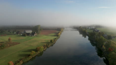 Morning-fog-hangs-over-a-smooth-river-and-peaceful-green-farms-along-the-banks