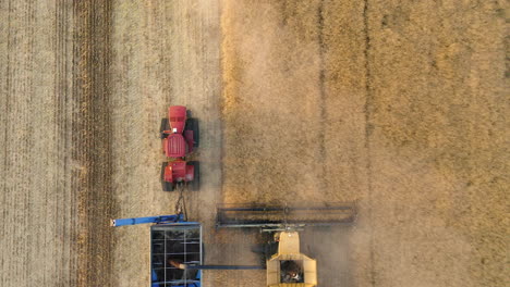 Top-down-of-combine-harvester-loading-grain-cart-behind-tractor-on-yellow-field