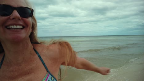 Closeup-of-laughing,-playful-mature-woman-in-bikini-and-sunglasses-turns-around-and-runs-on-the-beach