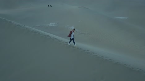 4K-Aerial-follow-shot-of-a-boy-walking-alone-in-a-barren-desert-on-a-large-sand-dune-during-a-windy-and-dry-day